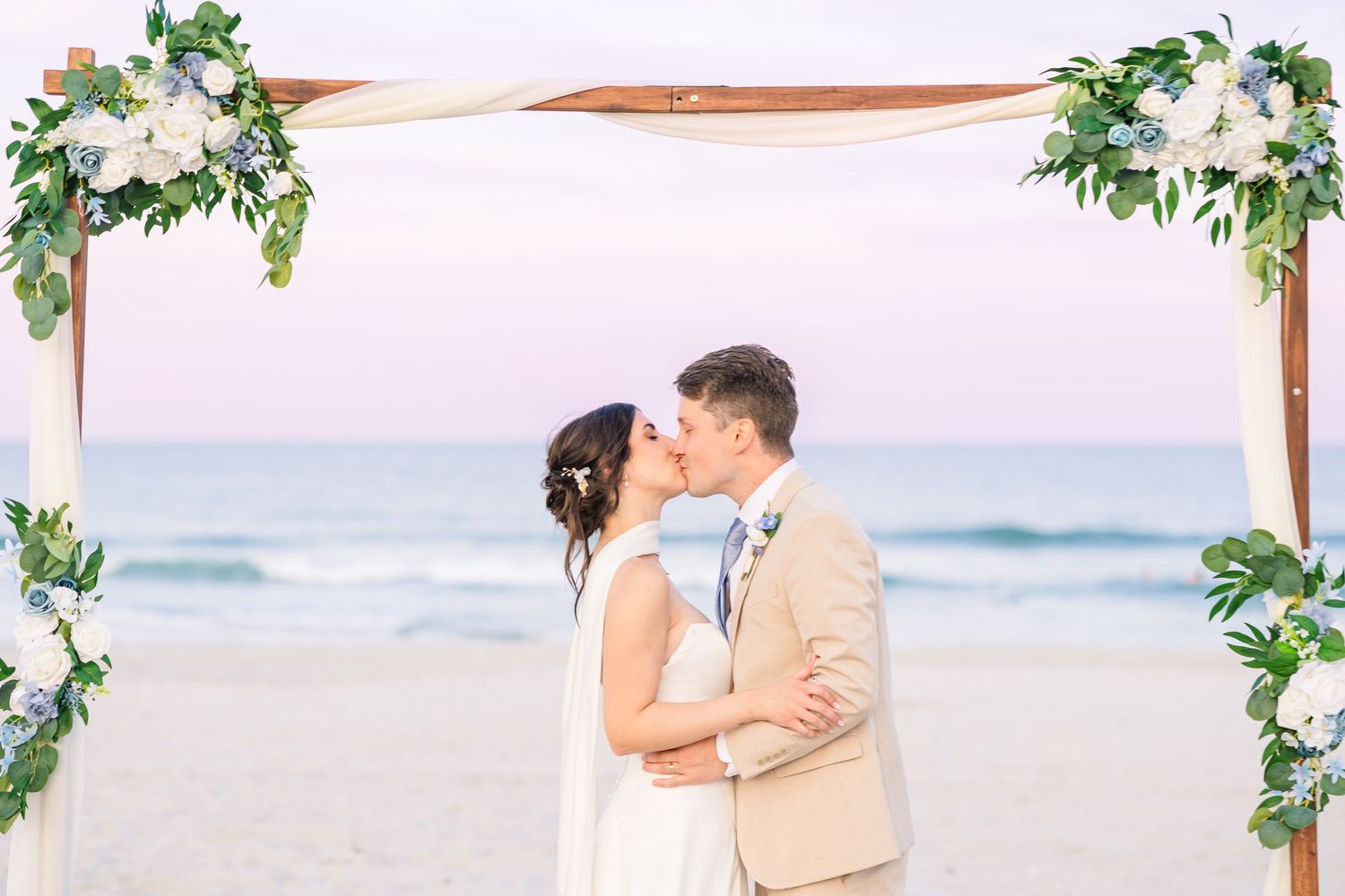 Newlyweds share first kiss under floral wooden arch on beach at sunset with pastel sky backdrop in Carolina Beach near Wilmington, NC.