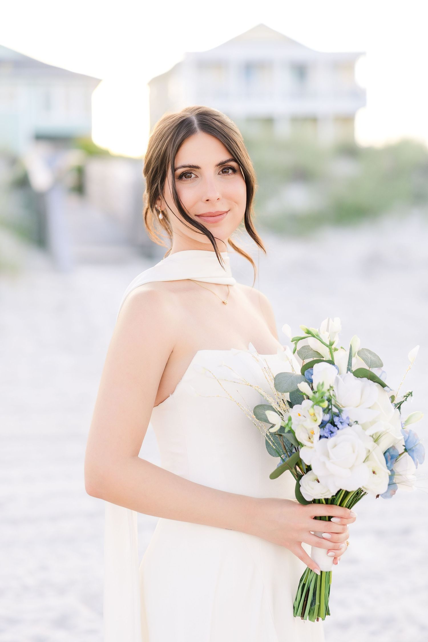 Elegant bride in white strapless gown holds blue and white floral bouquet on a sunlit sandy beach in Carolina Beach near Wilmington, NC.