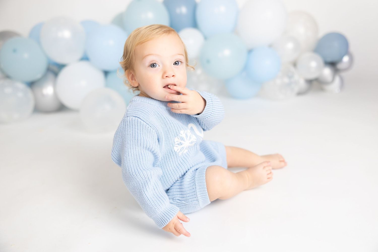 Baby in light blue outfit plays on white floor with balloon decoration.