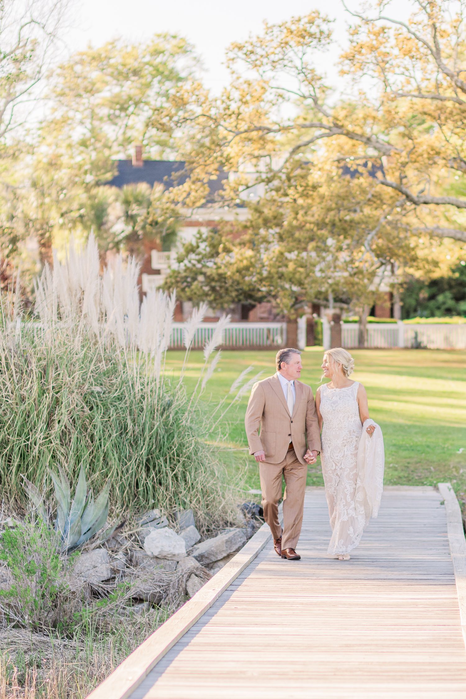 Couple walks along wooden boardwalk surrounded by tall grass and autumn trees.
