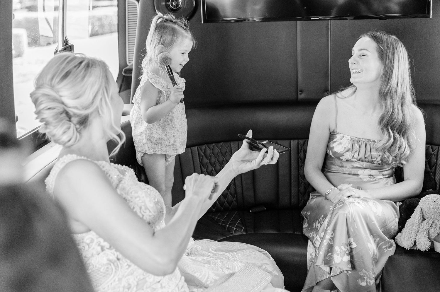 Women in formal attire share a candid moment inside a vehicle in black and white.