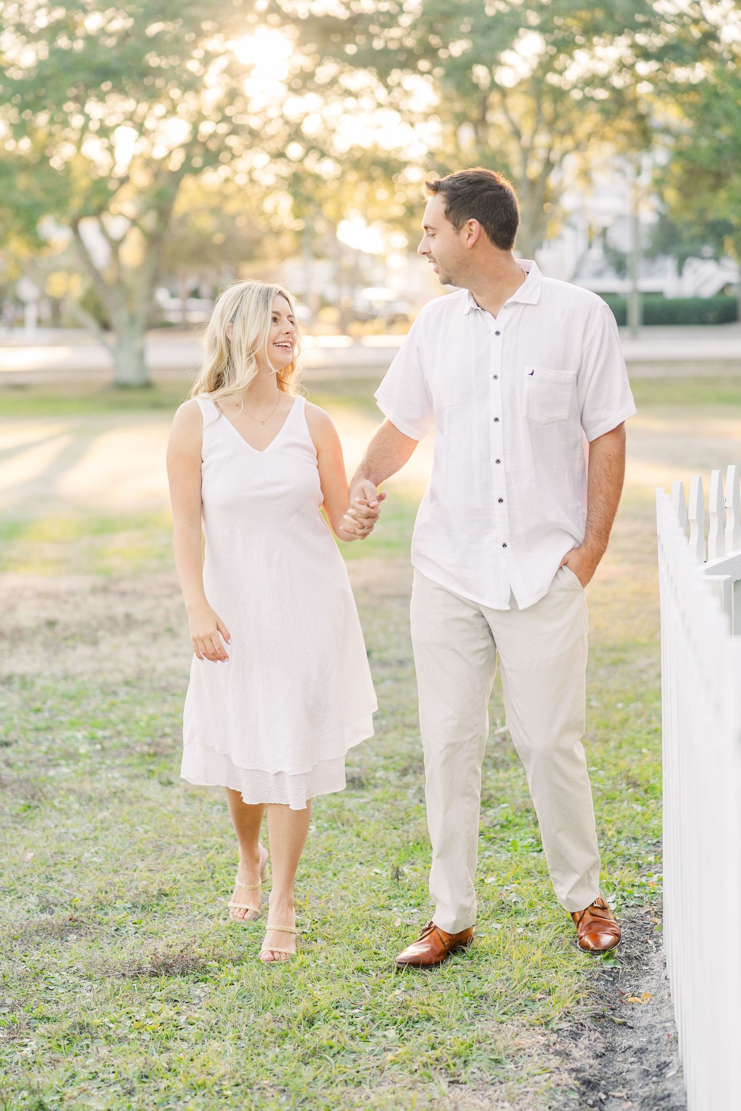 Couple in matching white outfits walking together through a park during golden hour.