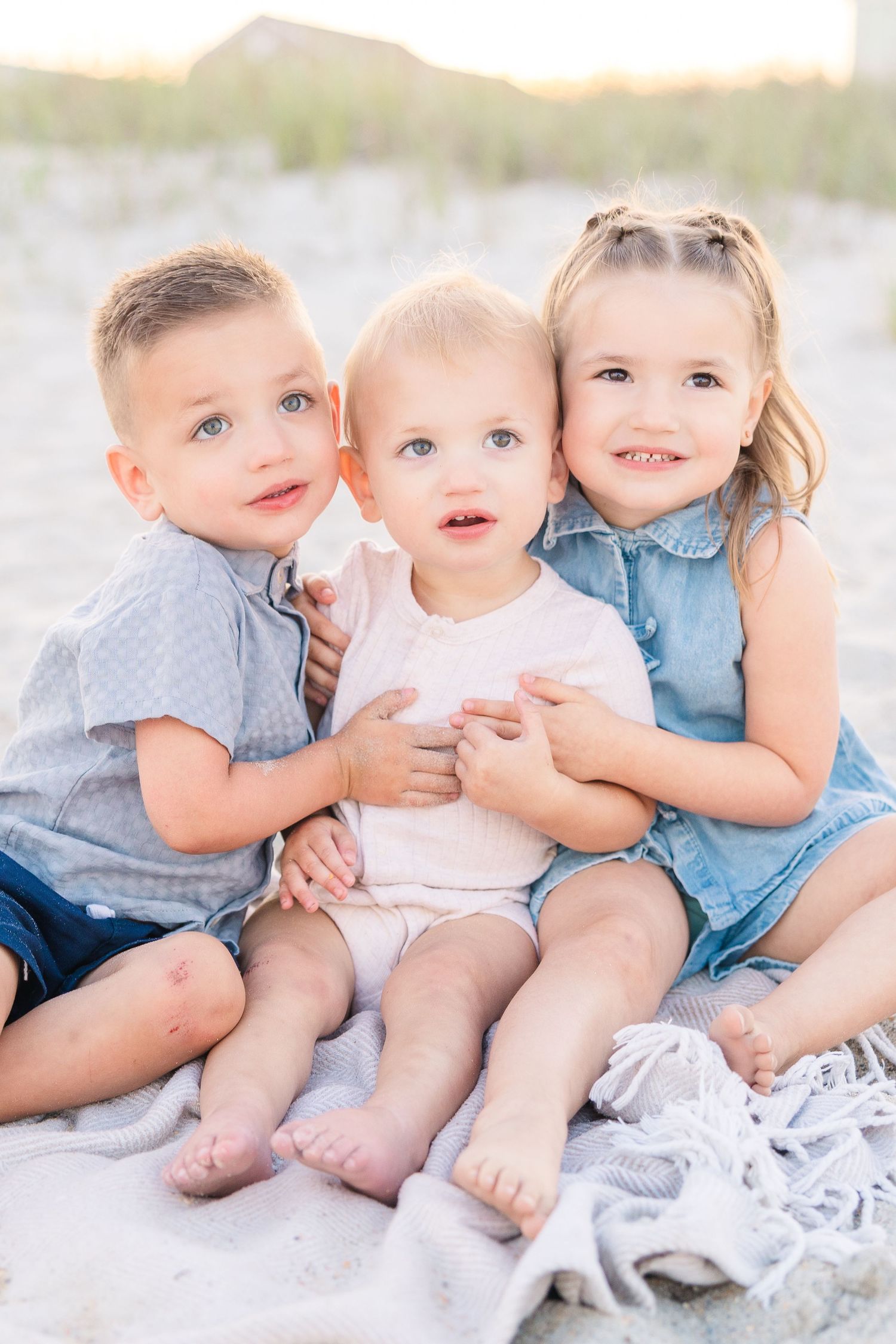 Three young siblings sit together on a blanket at the beach, smiling and hugging in a sweet family portrait.