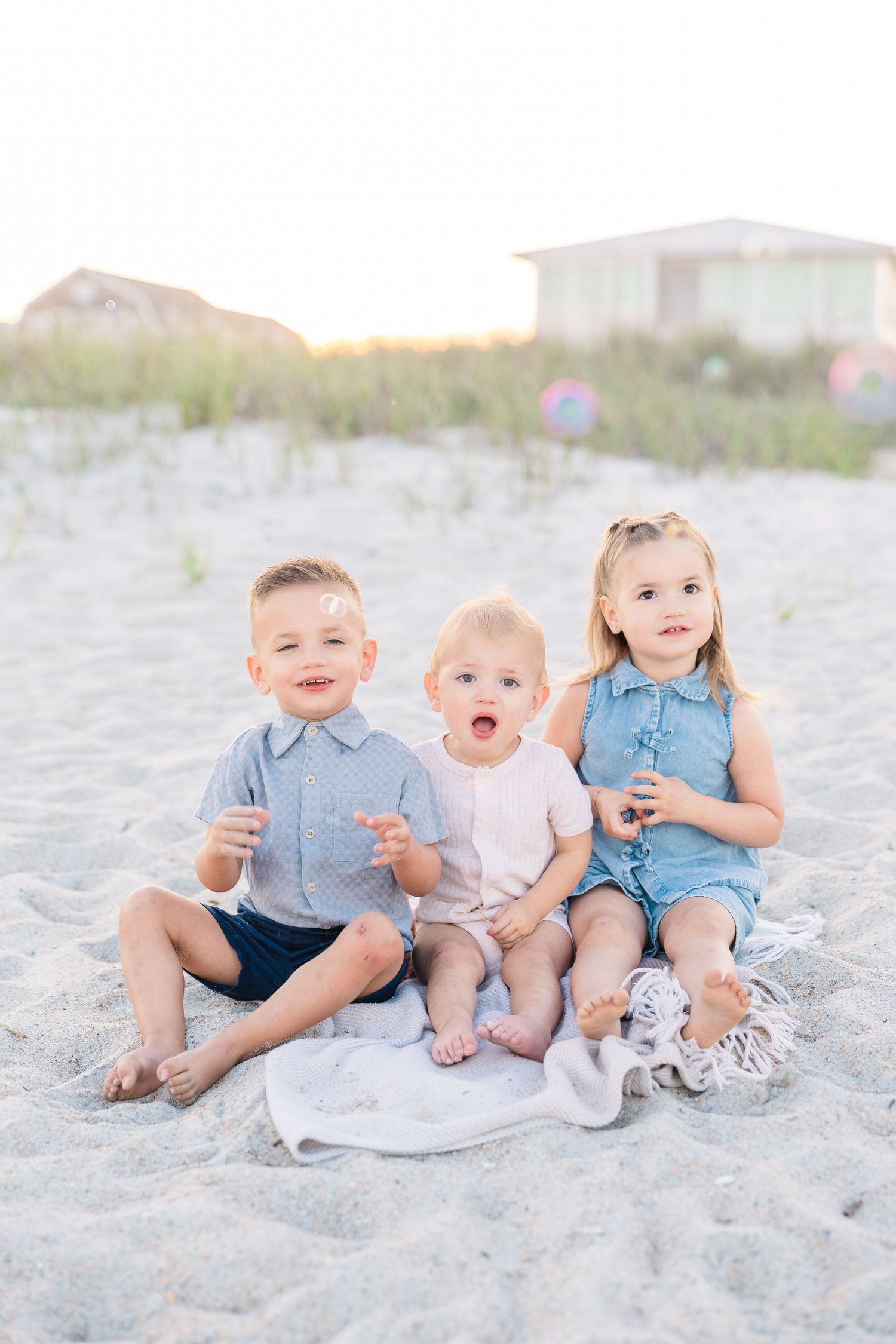 Three young children sit on sandy beach blowing bubbles, wearing pastel outfits with dunes and houses in background.
