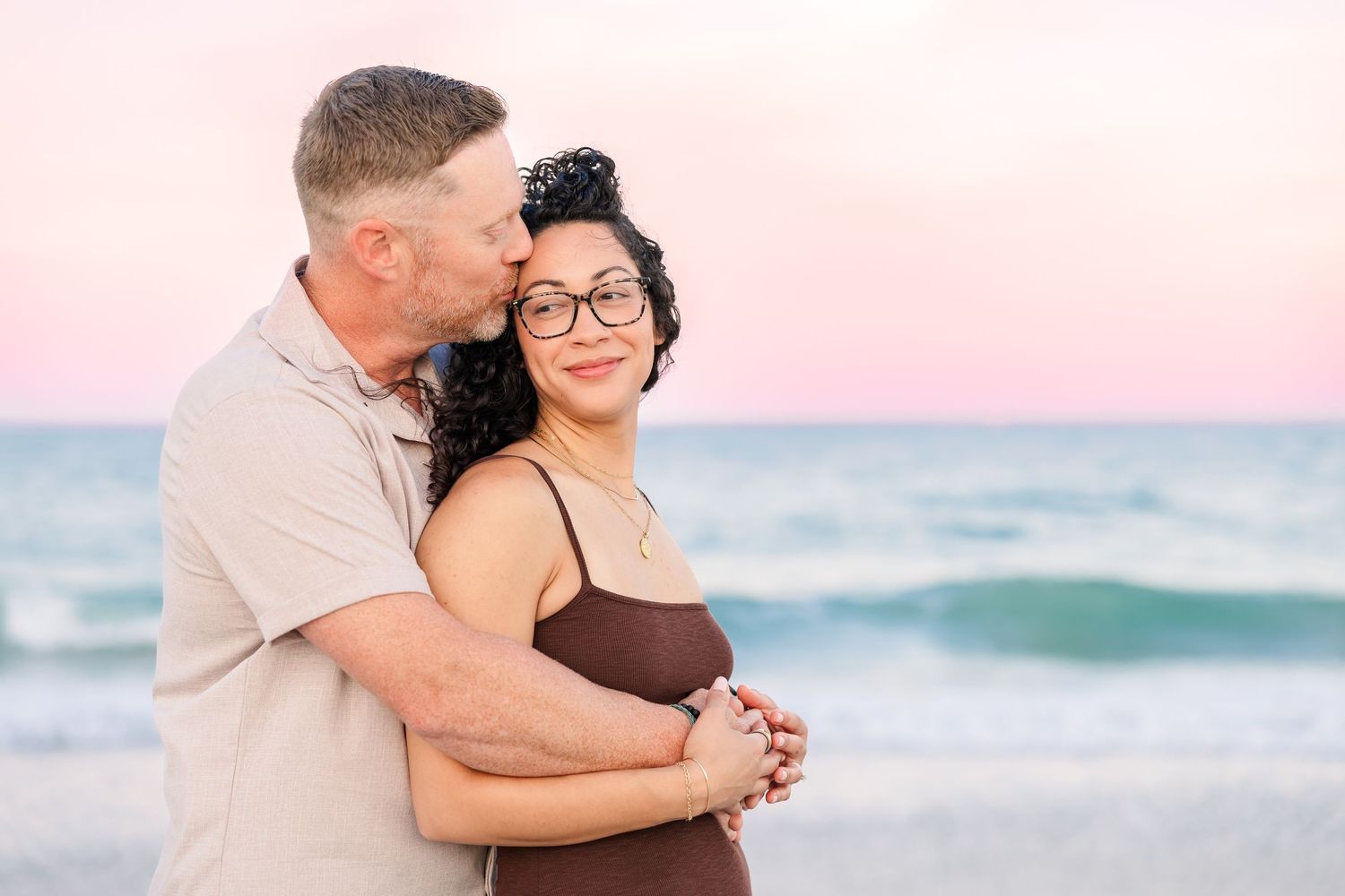 A couple embraces on a beach at sunset, man kissing woman's head, she smiles wearing glasses, pink sky backdrop.