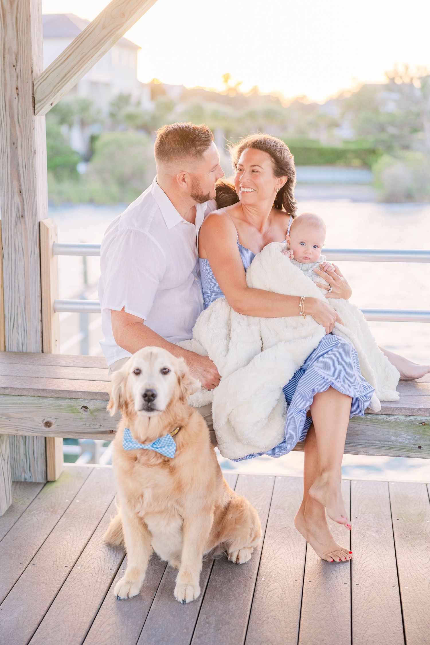 Family relaxes with their golden retriever on coastal boardwalk during sunset.