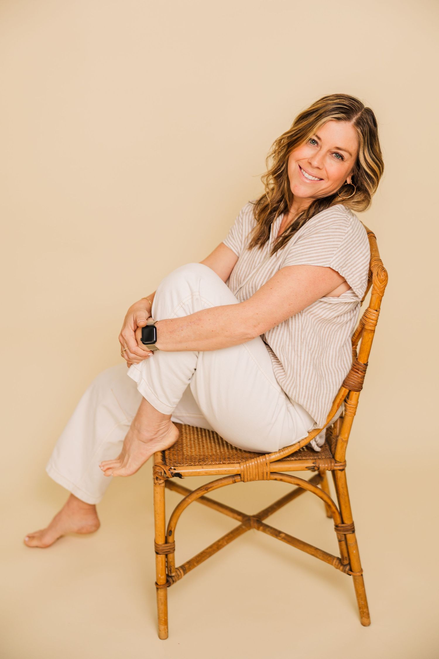 Casual portrait of someone sitting in bamboo chair wearing striped shirt and white pants.