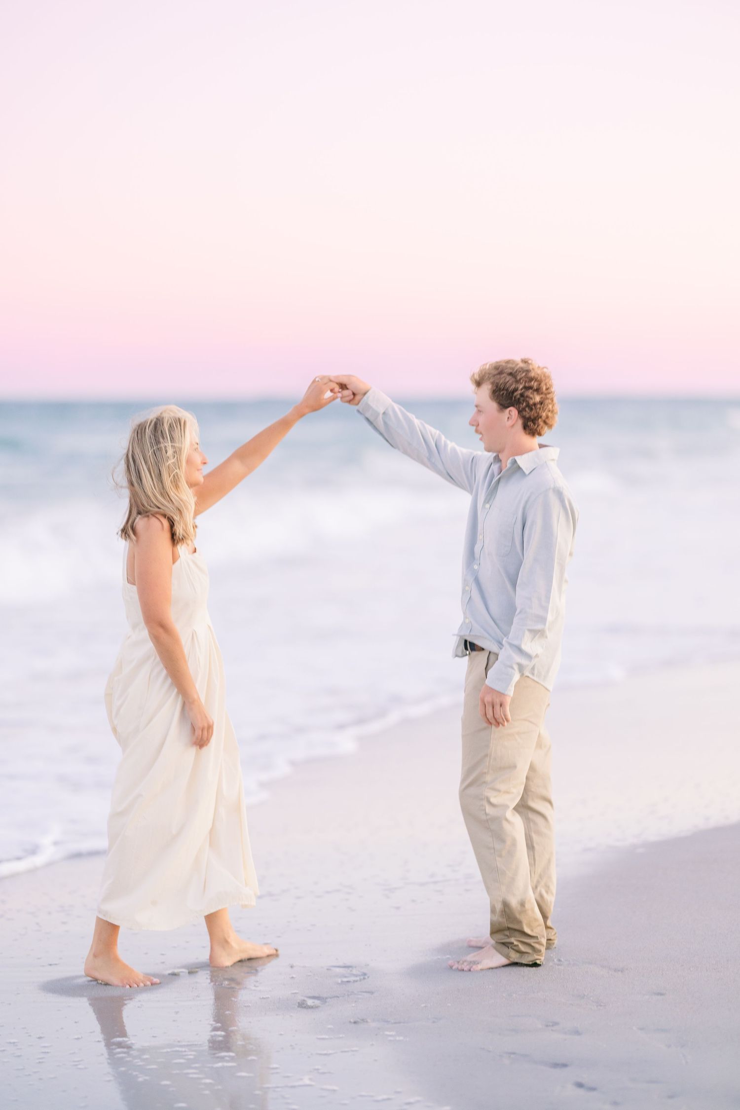 Two people dance and twirl together on a pristine beach with gentle waves and pink sky at dusk.