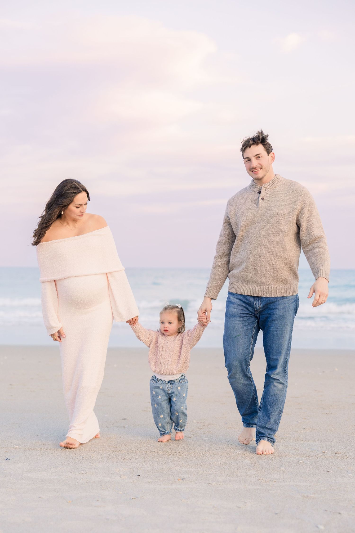 Family walking hand-in-hand on beach wearing neutral tones during sunset.