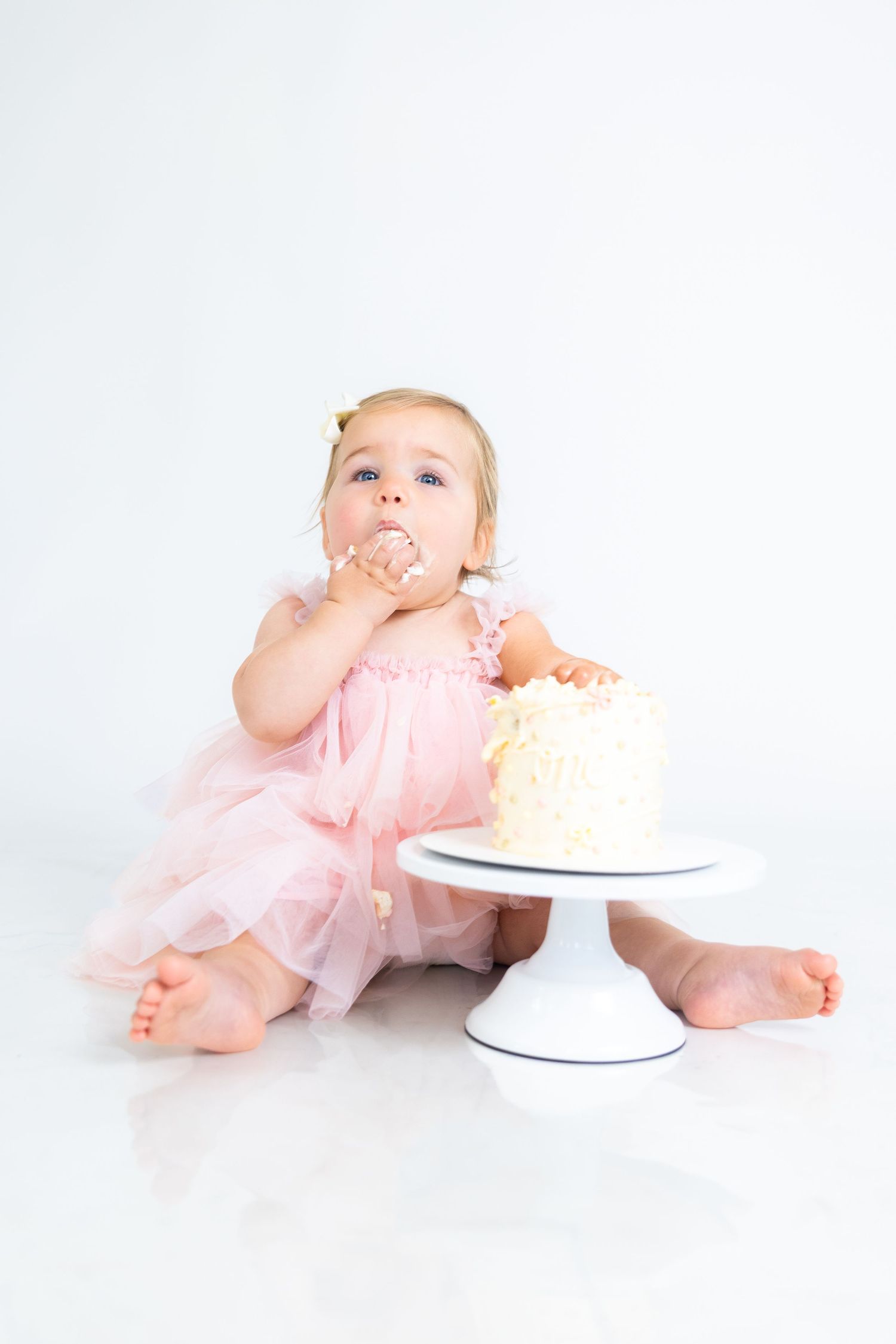Baby in pink dress lies on floor near cake stand during photo session.