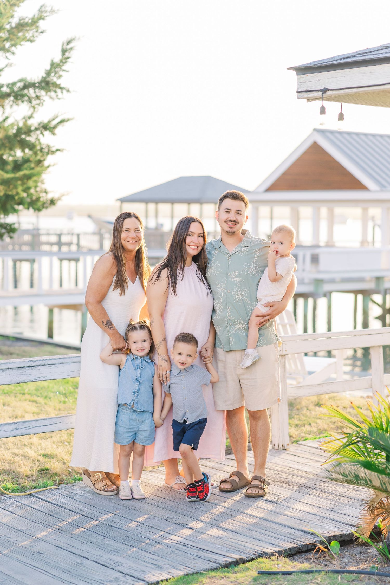 Extended family of six smiles together on waterfront dock at golden hour near a gazebo.