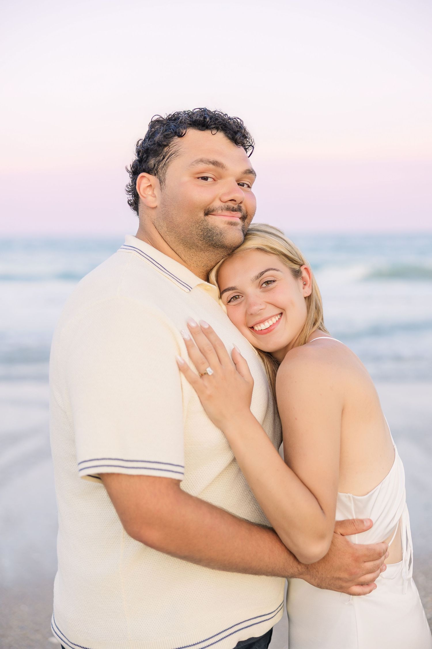 Couple embracing on beach at sunset, woman showing engagement ring, smiling at camera with ocean waves behind them.
