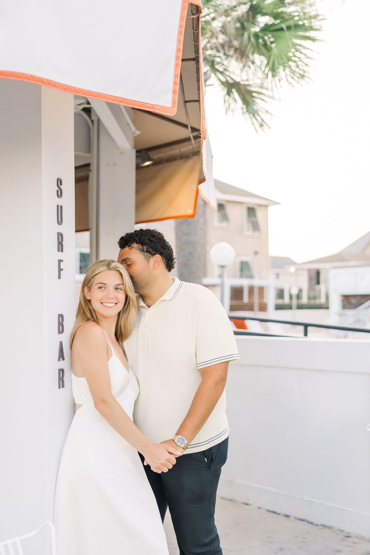 A smiling couple holds hands outside a Surf Bar, with the man kissing the woman's cheek near a palm tree.