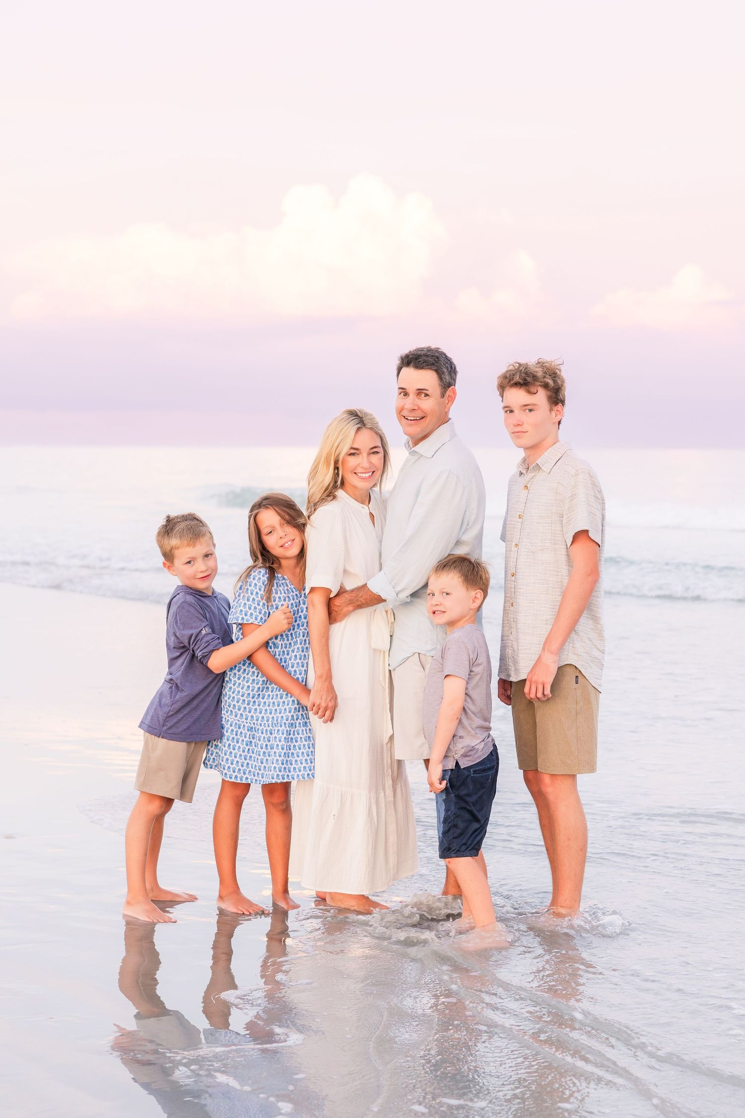 A family stands together at the edge of the ocean during sunset wearing coordinated white and blue outfits.