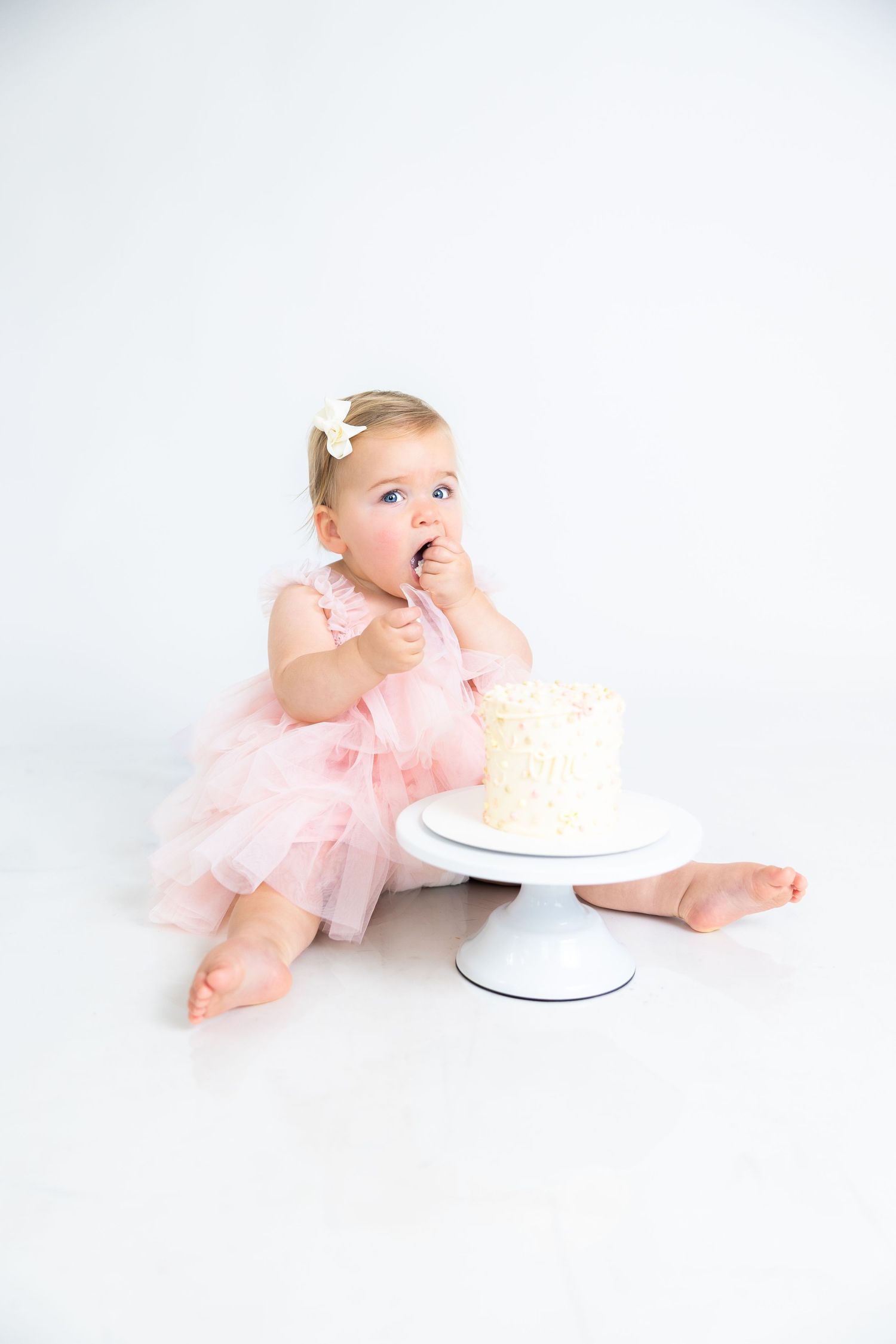 Baby in pink dress reaching for white cake on cake stand.