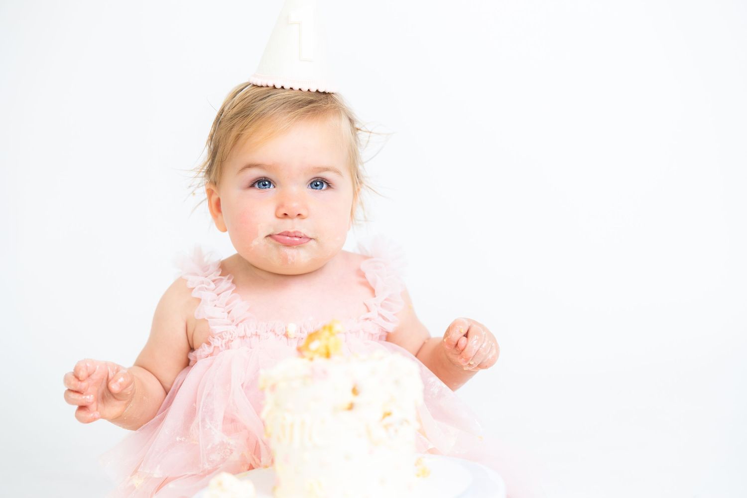 Baby in pink ruffled dress and party hat sitting next to white birthday cake.