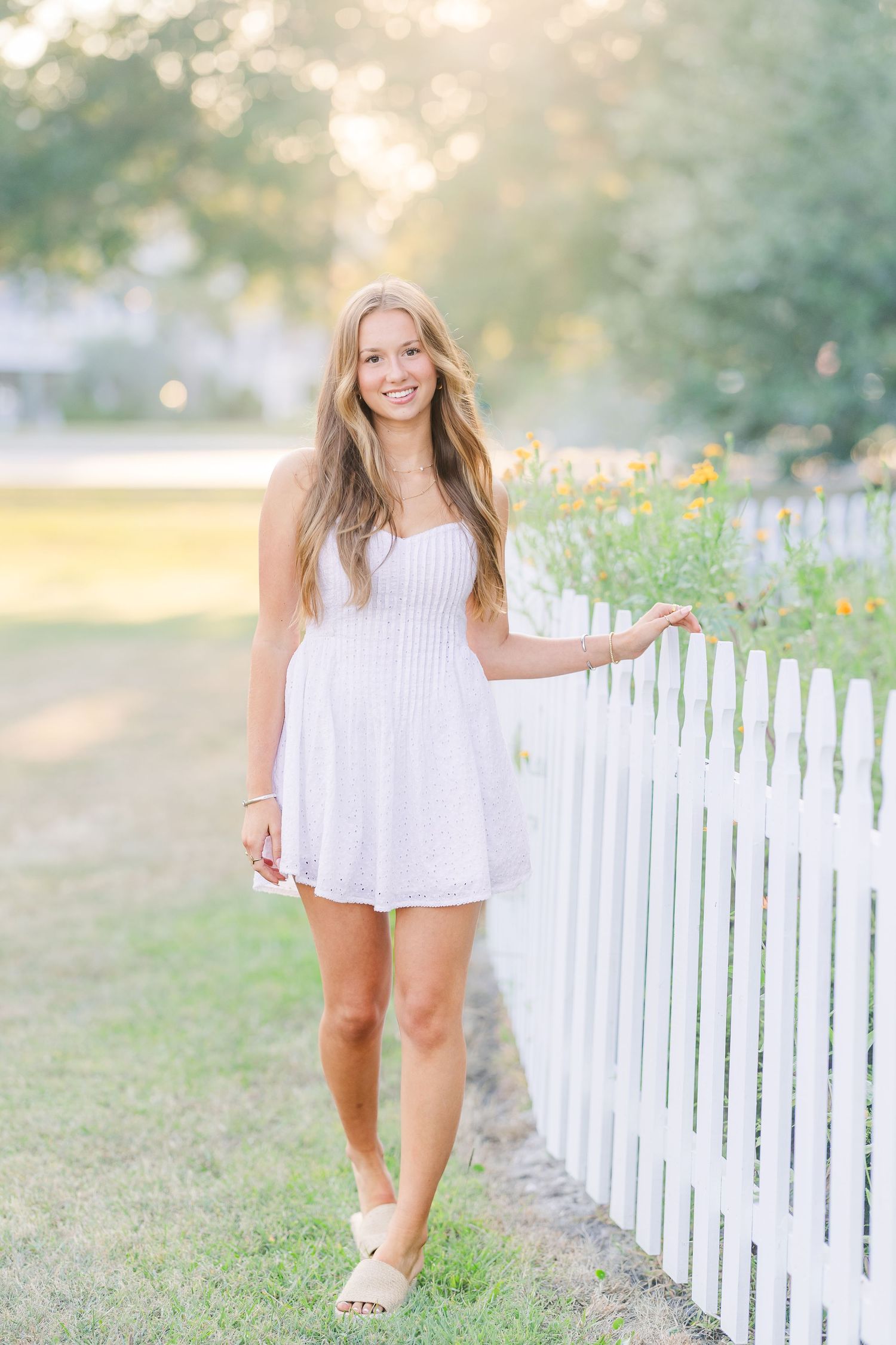 Person in white dress poses playfully by white picket fence in garden setting.