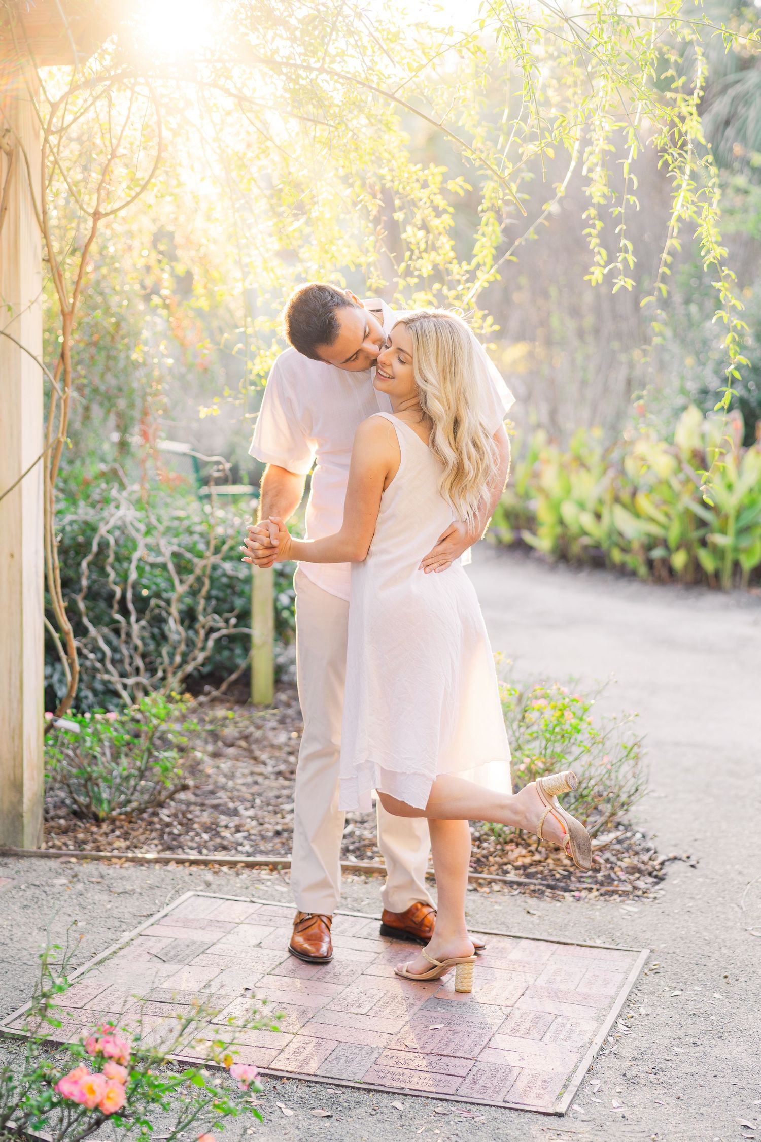 A romantic embrace in a sunlit garden path with flowering plants and warm golden light.