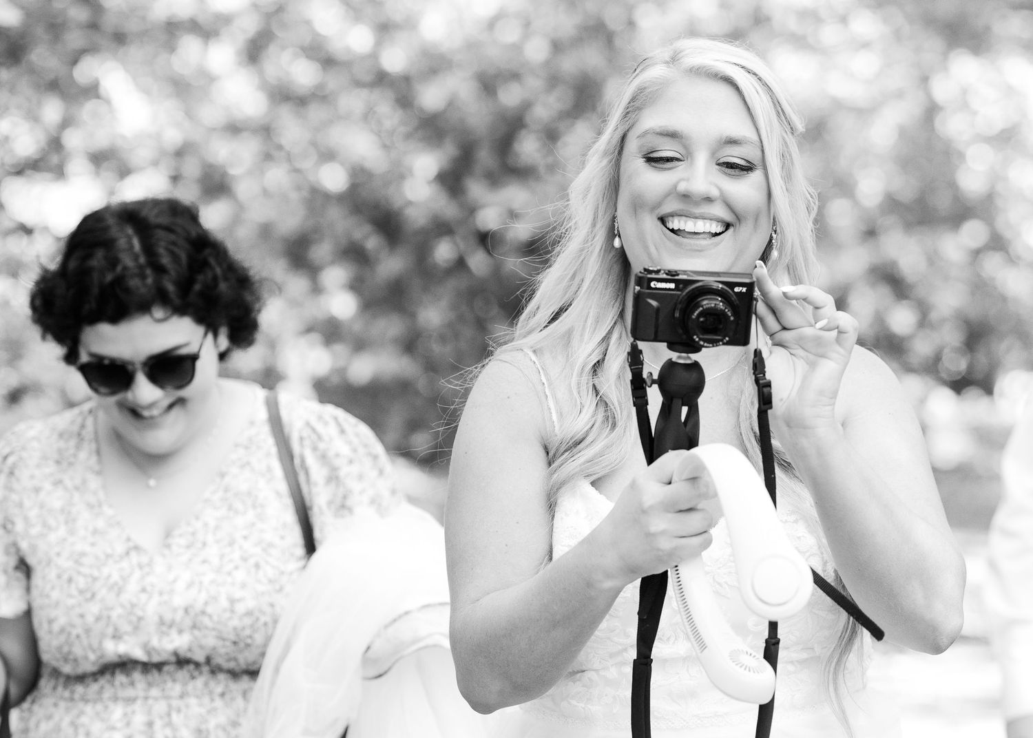 A smiling blonde woman holds a camera outdoors in black and white photo, with a brunette woman in sunglasses beside her.