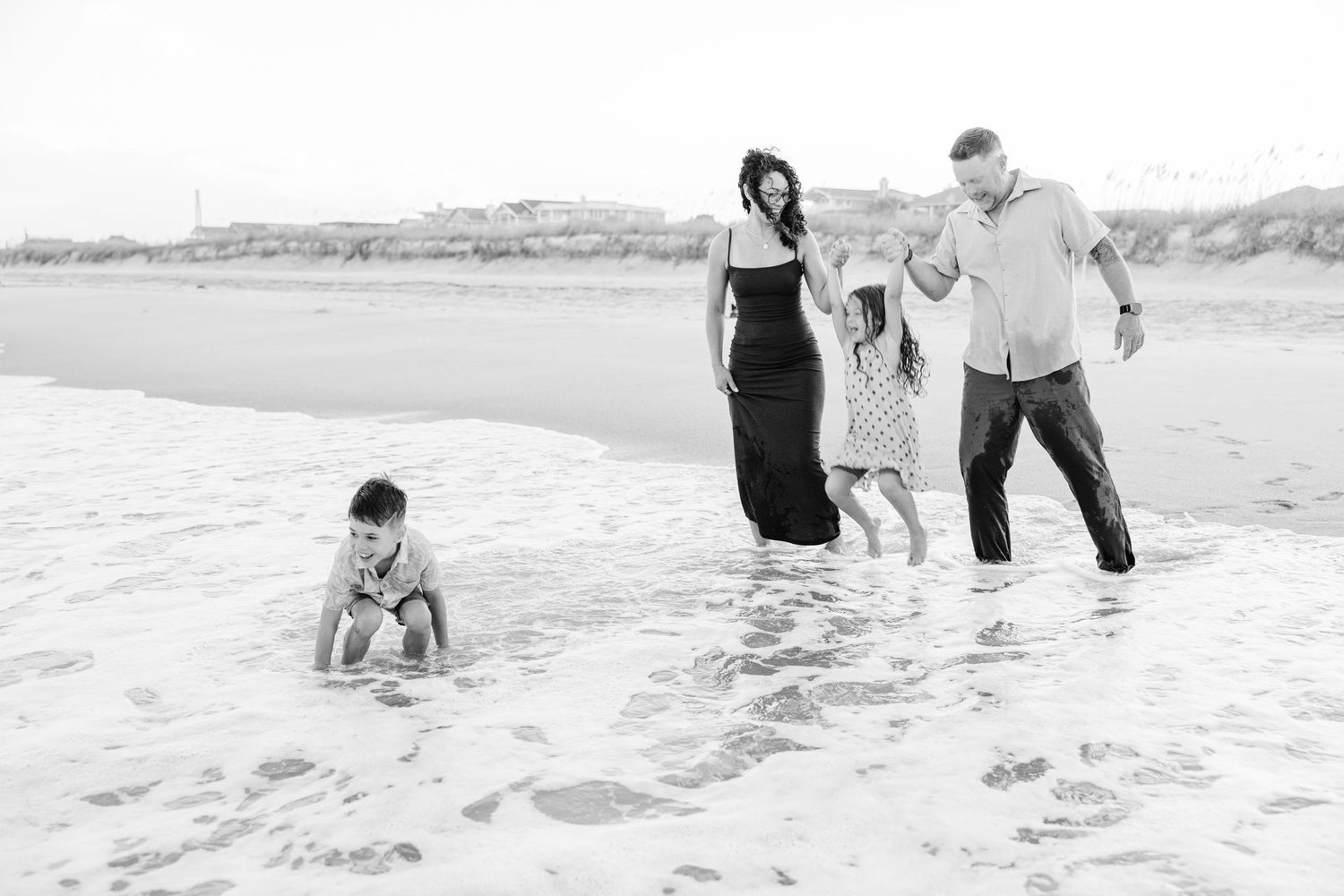 Black and white photo of a family of four playing in ocean waves on a sandy beach together.