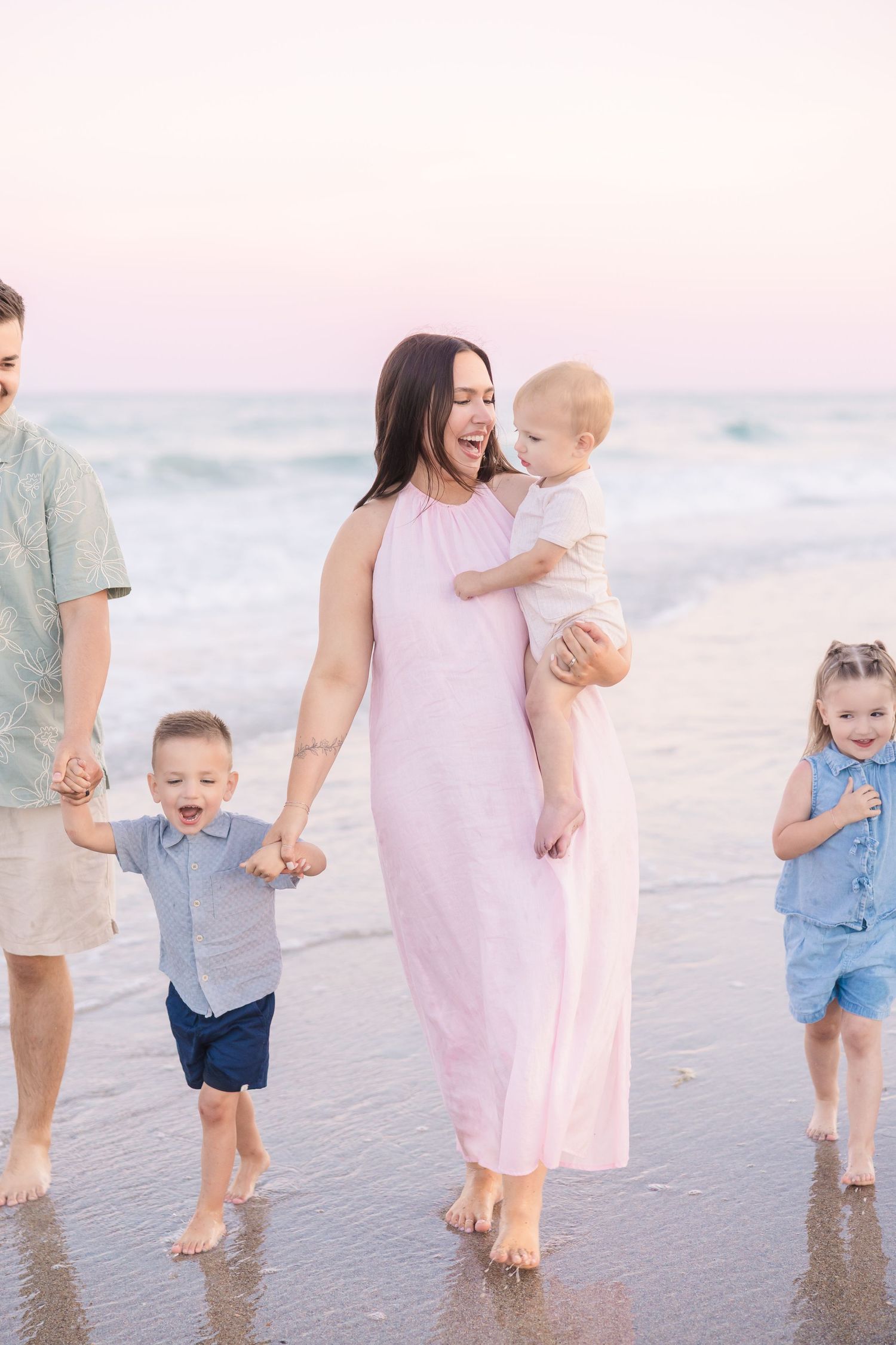 Happy family of five walking barefoot on beach shoreline at dusk, mother in pink maxi dress holding baby.
