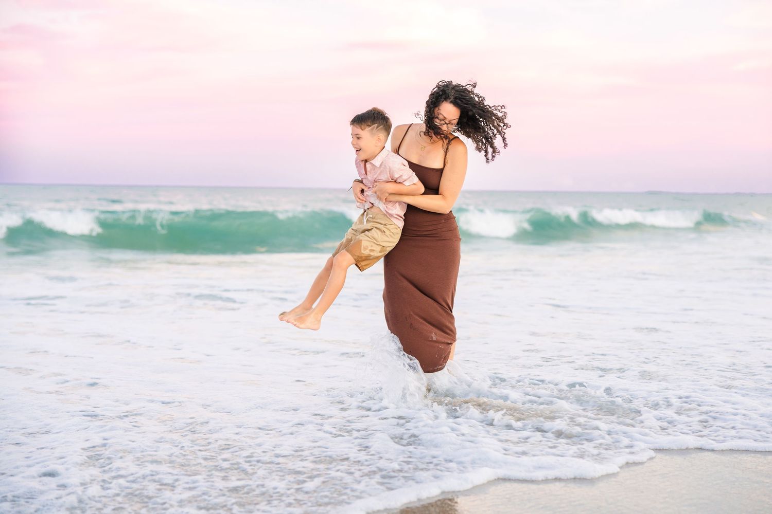 A mother in a brown dress joyfully carries her son through ocean waves at the beach.