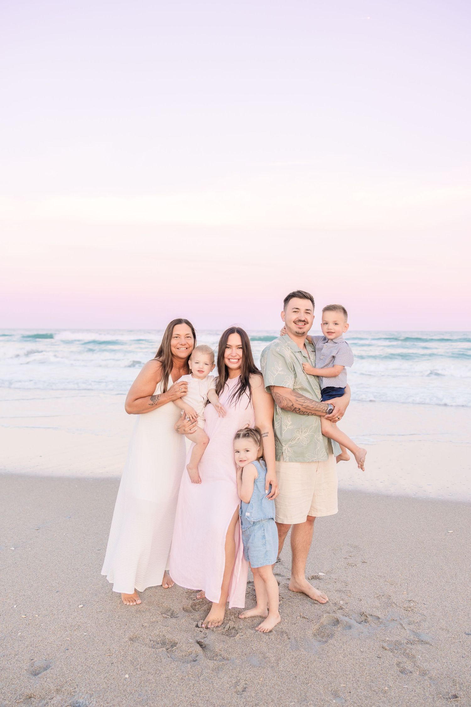 Extended family of six smiles together on beach at sunset, adults holding young children by the ocean.
