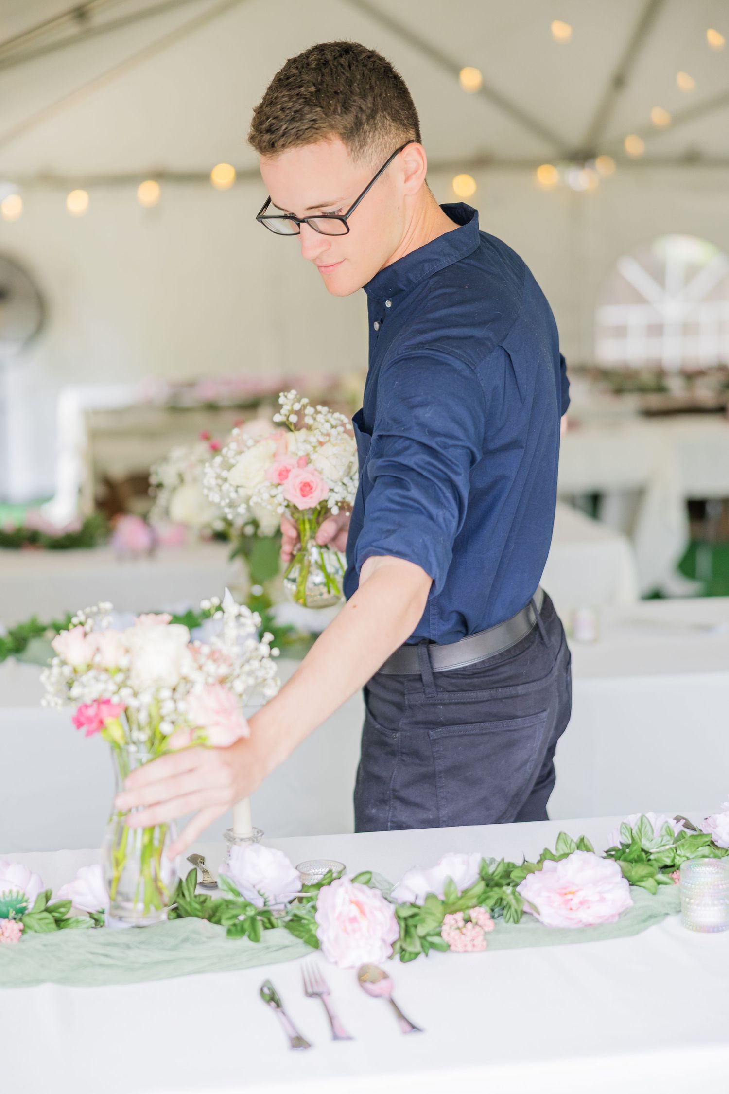 Wedding florist arranging flowers in glass vases under tent with string lights wedding groom bride wilmington North Carolina light and airy