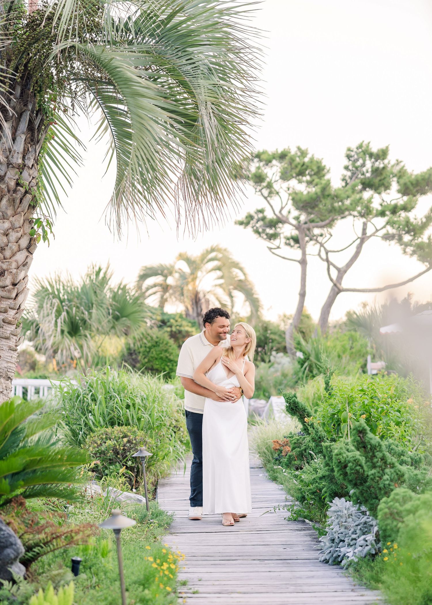 Couple embracing on a wooden boardwalk surrounded by lush tropical gardens and palm trees at sunset.