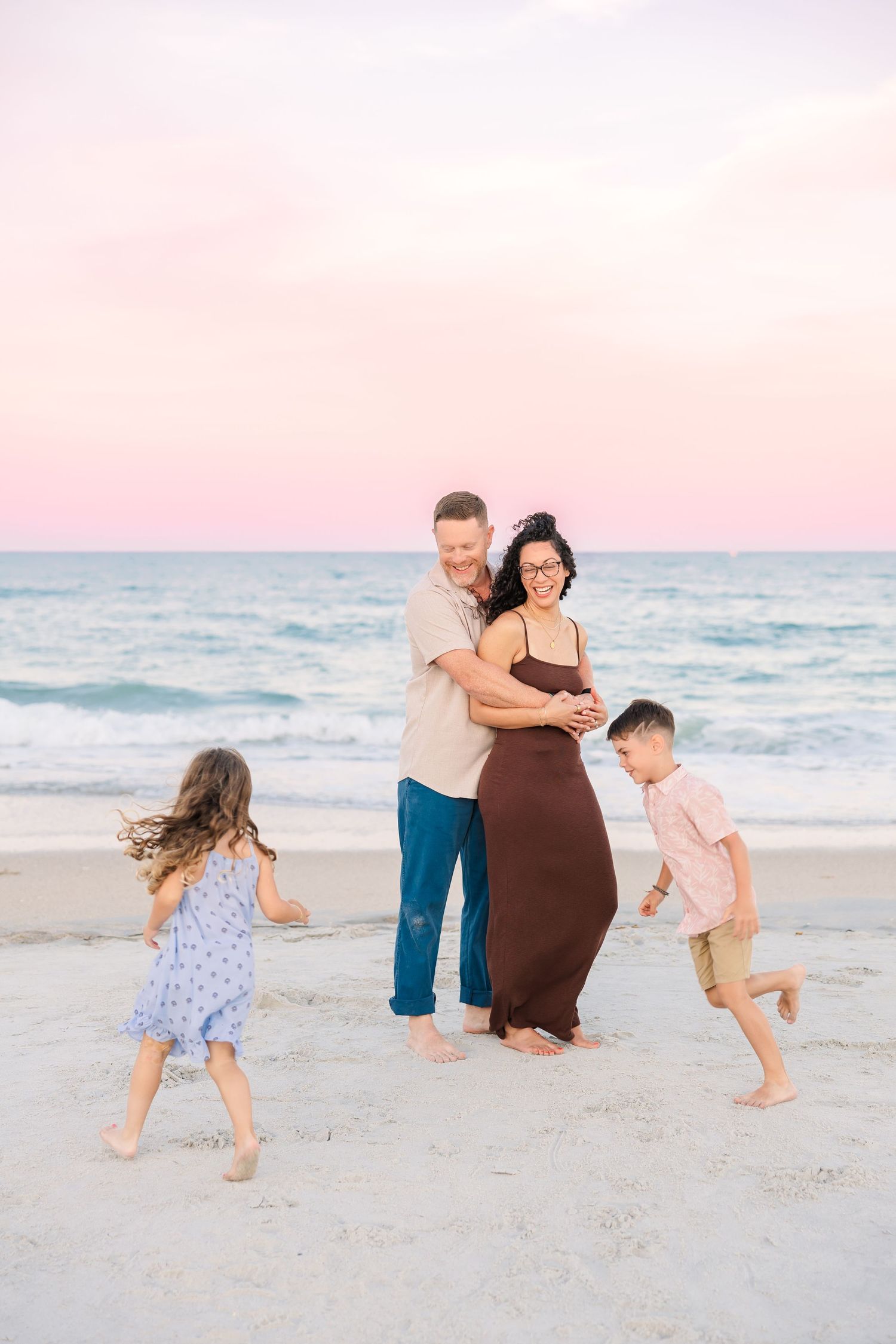 Family of four enjoys a playful sunset moment on the beach with pink sky and ocean waves behind them.