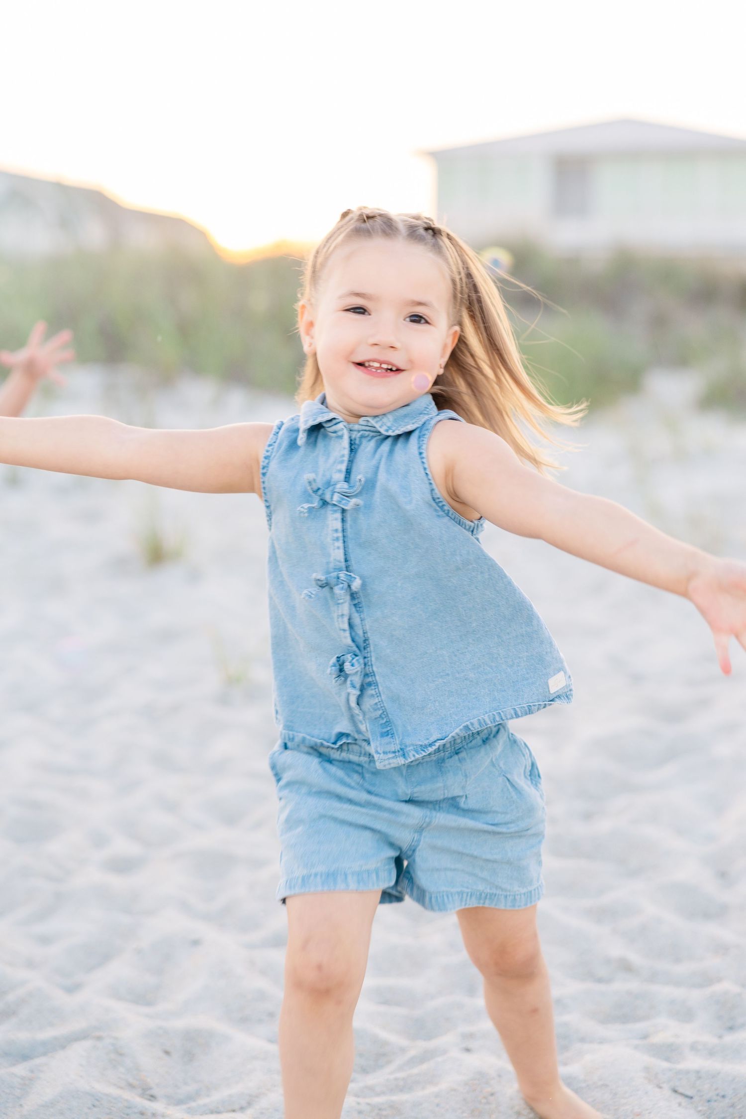 Joyful little girl in denim romper twirling on sandy beach at golden hour sunset.