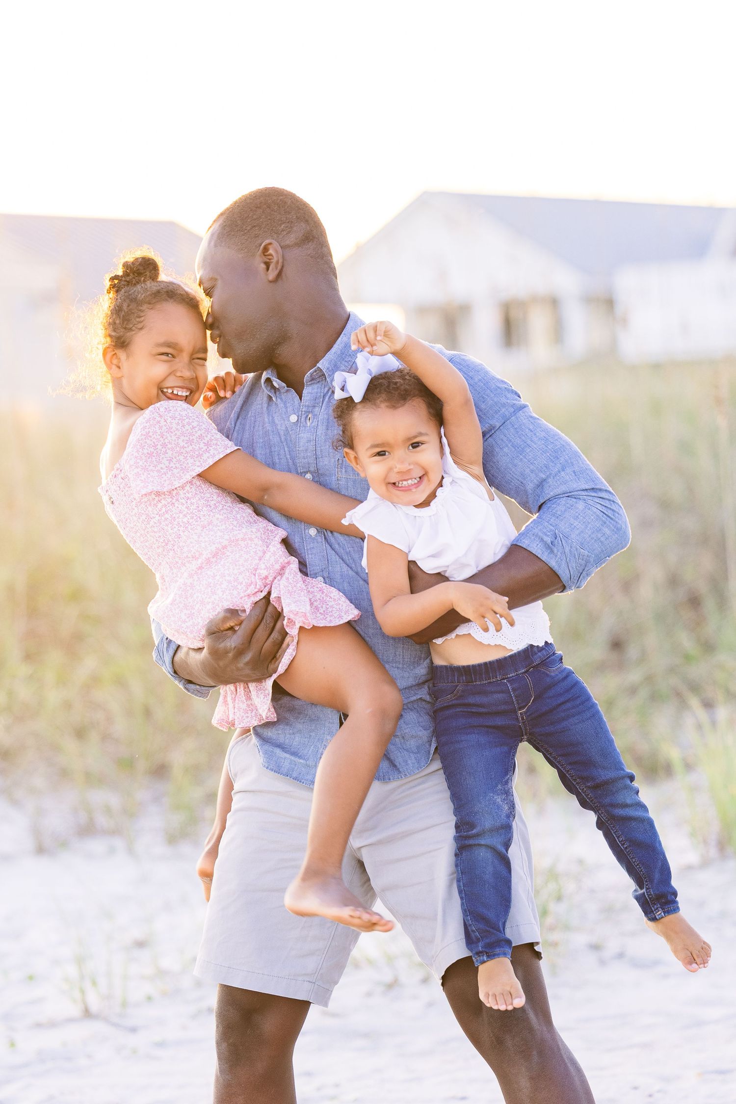 Family enjoying playful moments together on a sunny beach with soft lighting and sand dunes in the background.