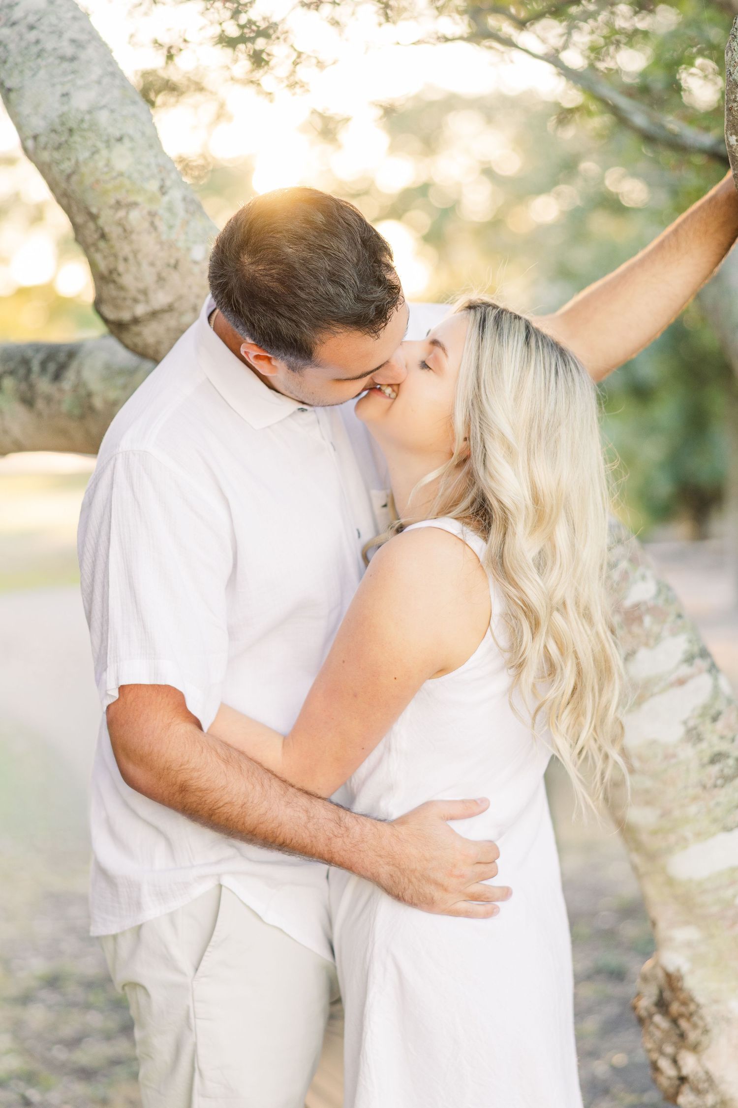Romantic couple in white embracing under trees during sunset photoshoot.