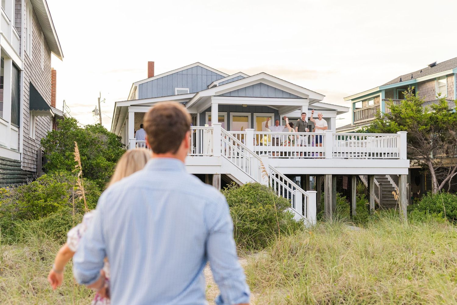 A couple walks towards a charming beach house with white railings and coastal vegetation.