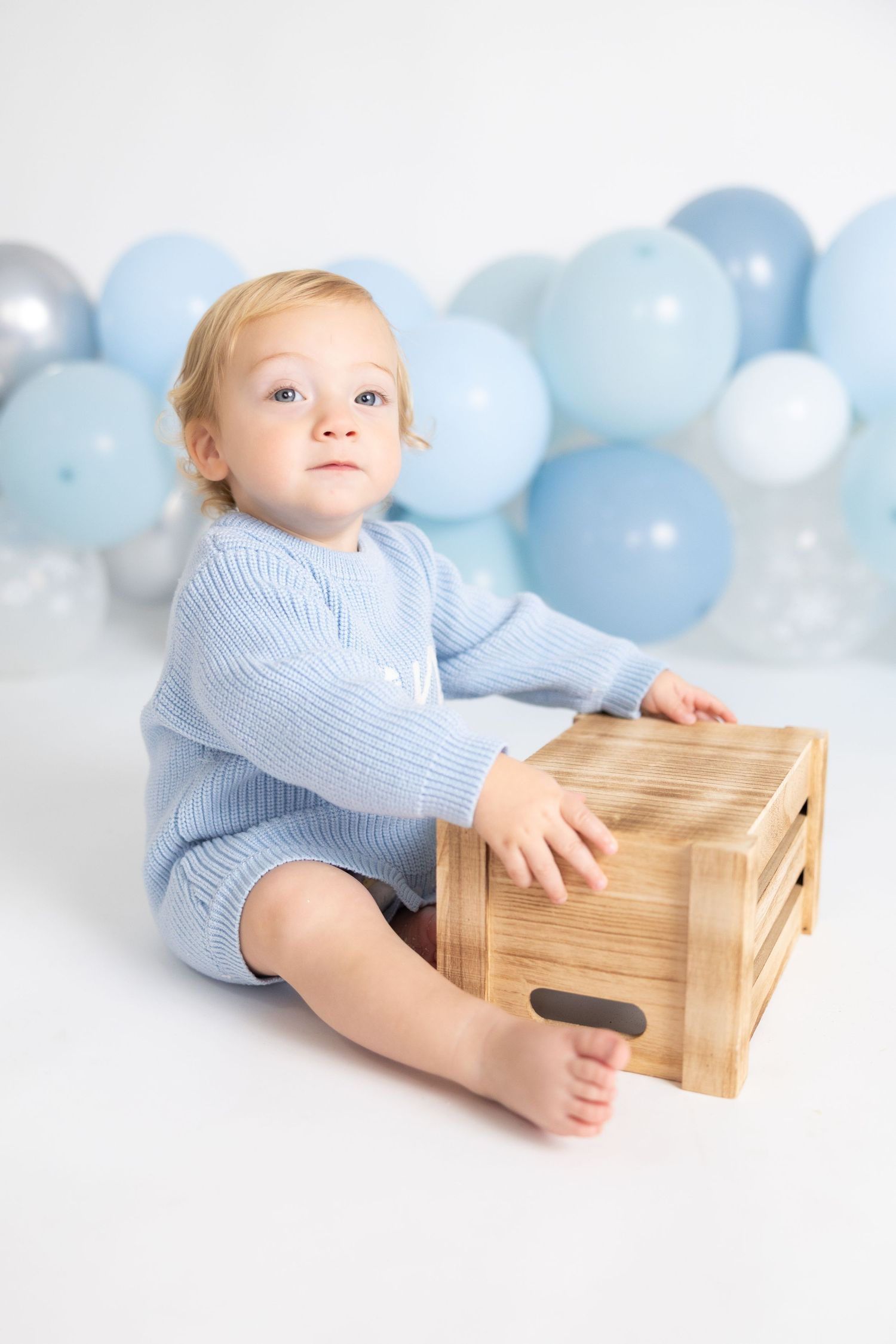 Baby in light blue outfit sits with wooden crate against backdrop of pastel blue and white balloons.