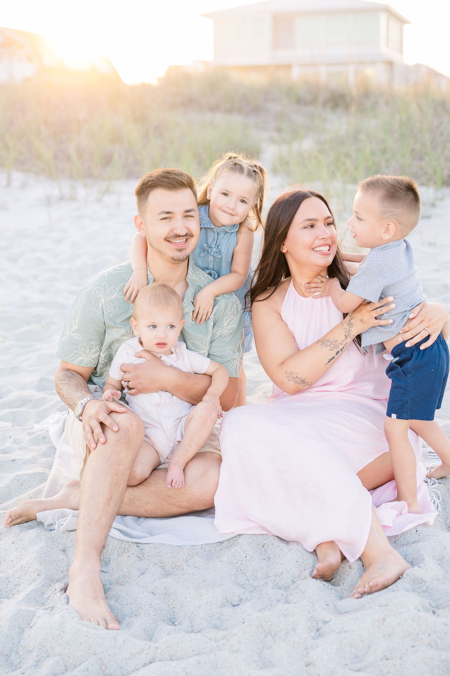 A happy family of five poses on a sandy beach at golden hour, with parents and three young children smiling warmly.