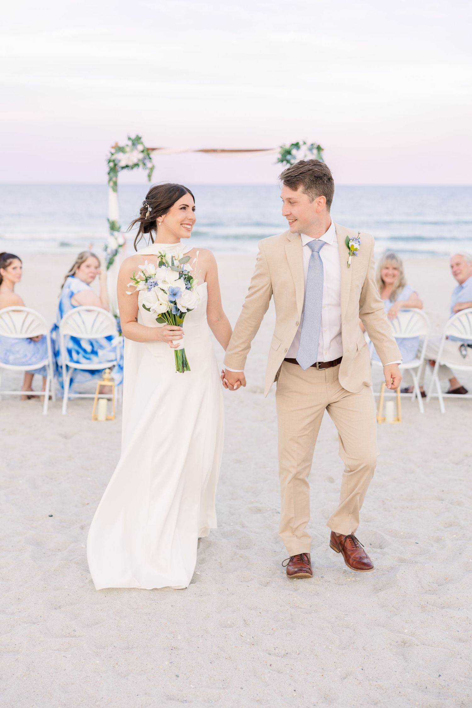 Bride and groom hold hands walking on beach after wedding ceremony with ocean backdrop at sunset in Carolina Beach near Wilmington, NC.