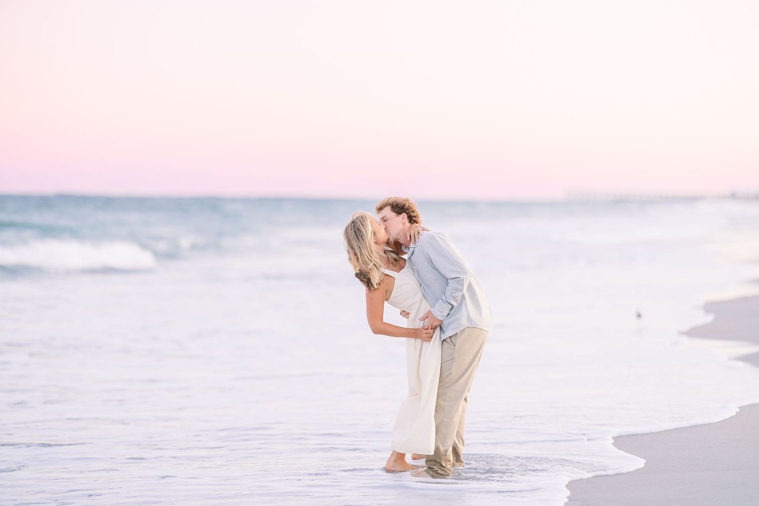 A couple in white and beige outfits dance playfully on a beach at sunset with soft pastel colors in the background.