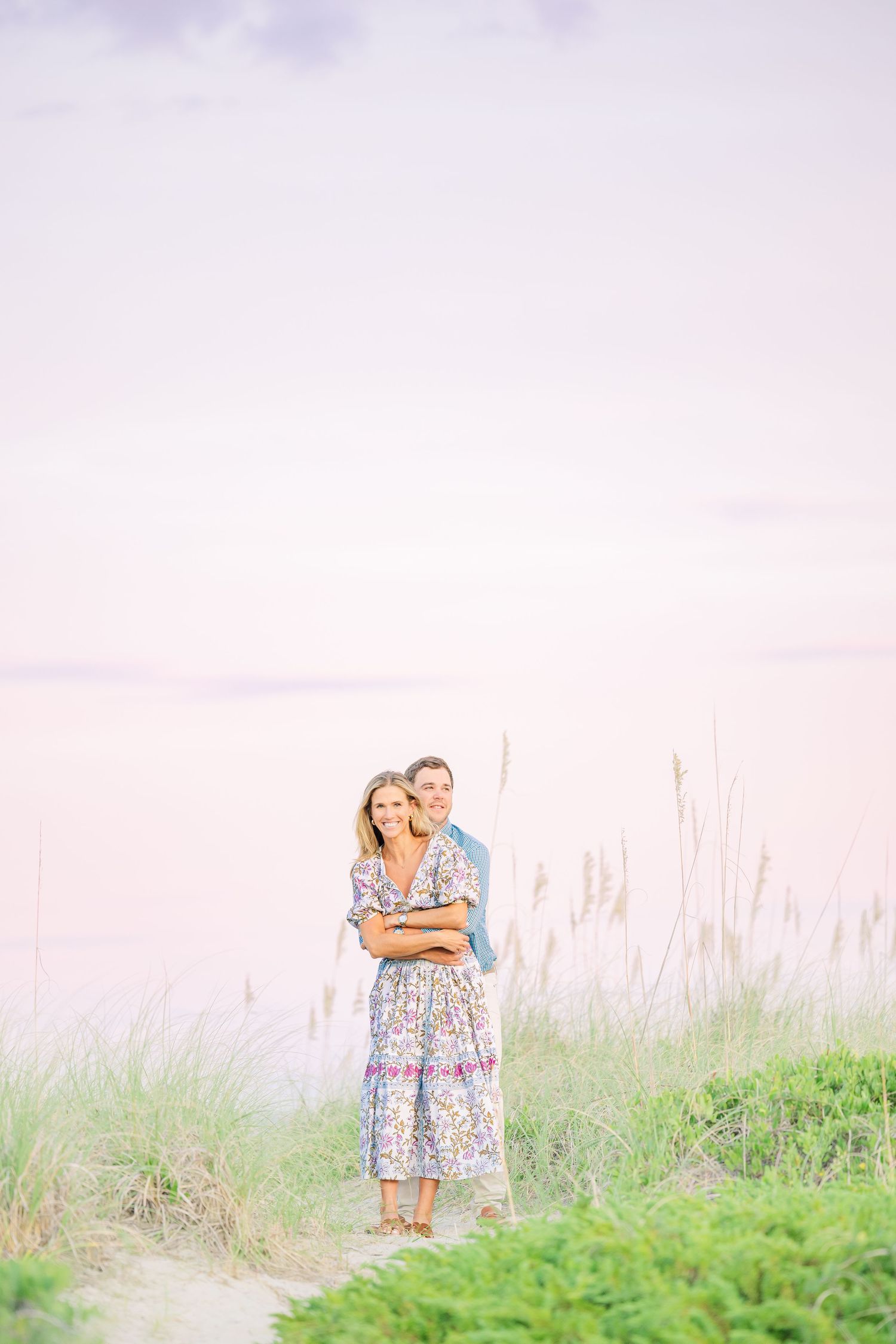 Romantic couple embraces on beach path with sea grass and pastel sky in background.