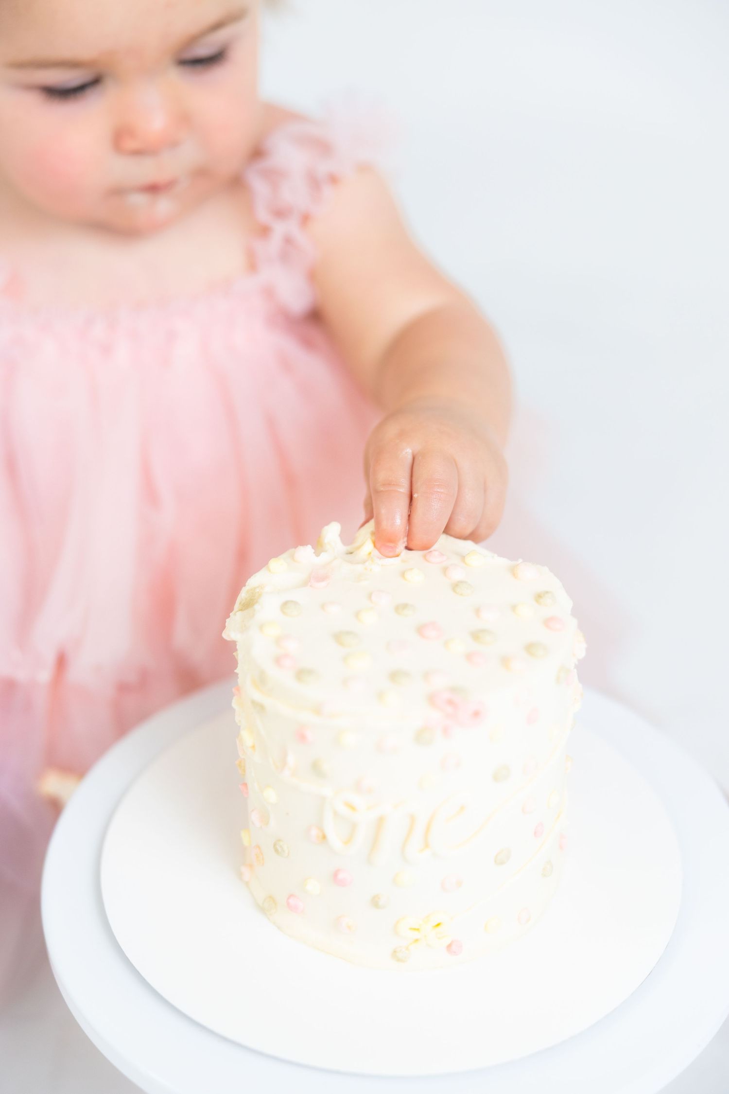 Small hands reach for white frosted cake on cake stand against white backdrop.