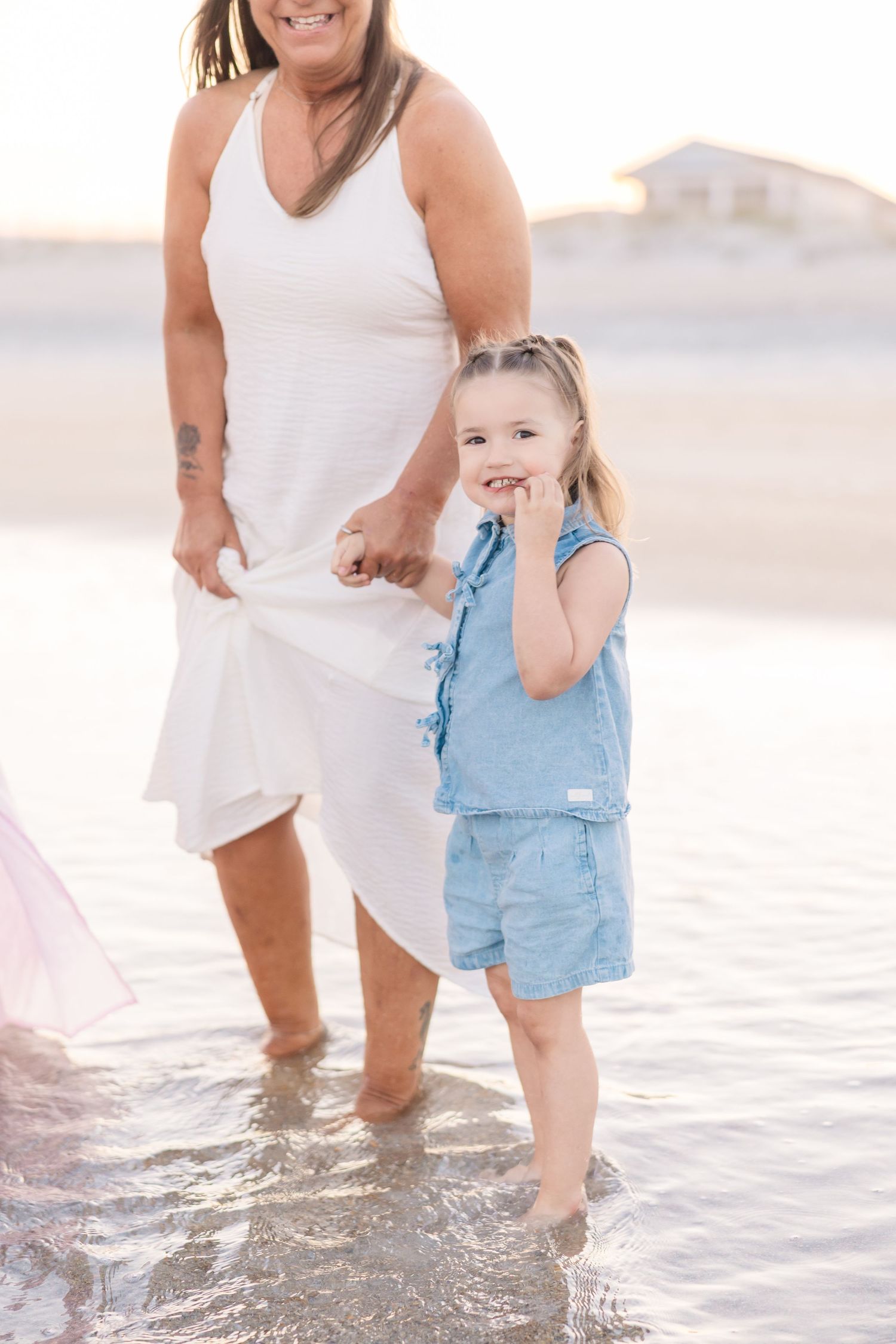 Young girl in denim romper smiling while standing in ocean water beside a woman in a white dress.