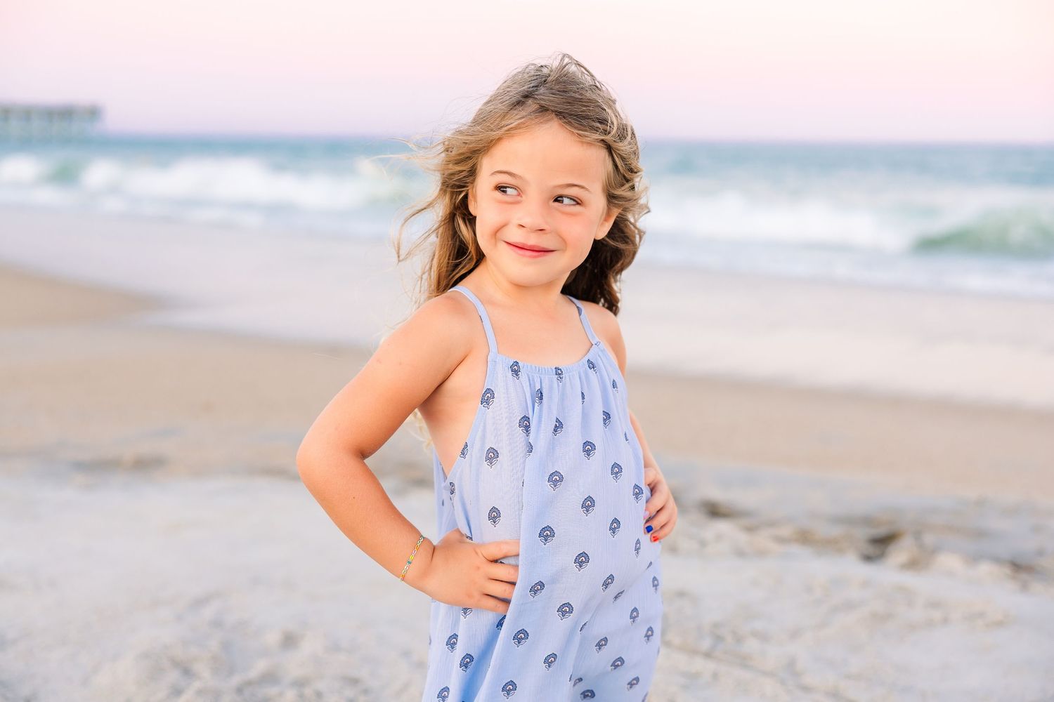 Young girl in a blue sundress posing confidently on a beach with ocean waves behind her.