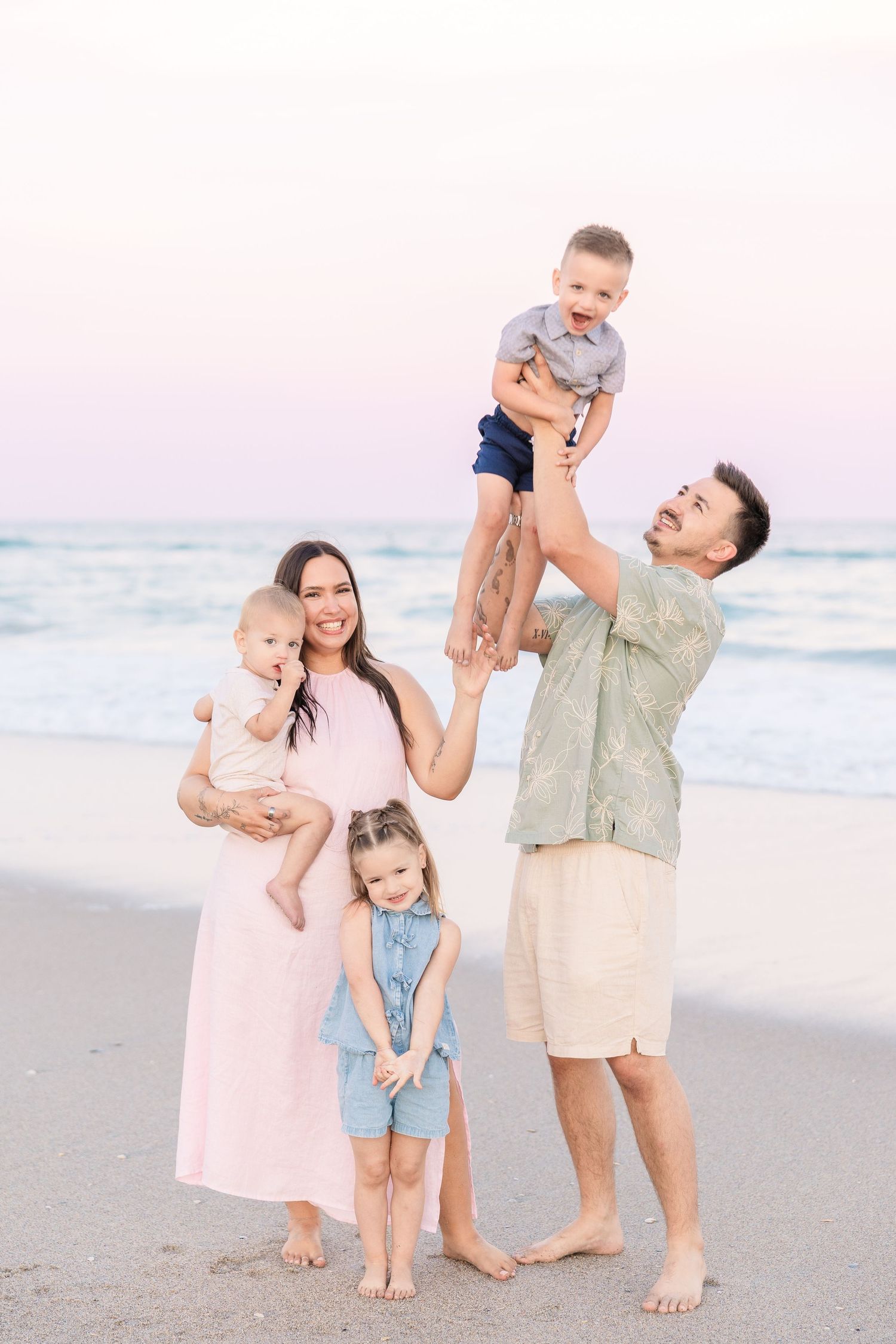 Father lifts laughing toddler overhead on beach while mother holds baby and daughter stands nearby.