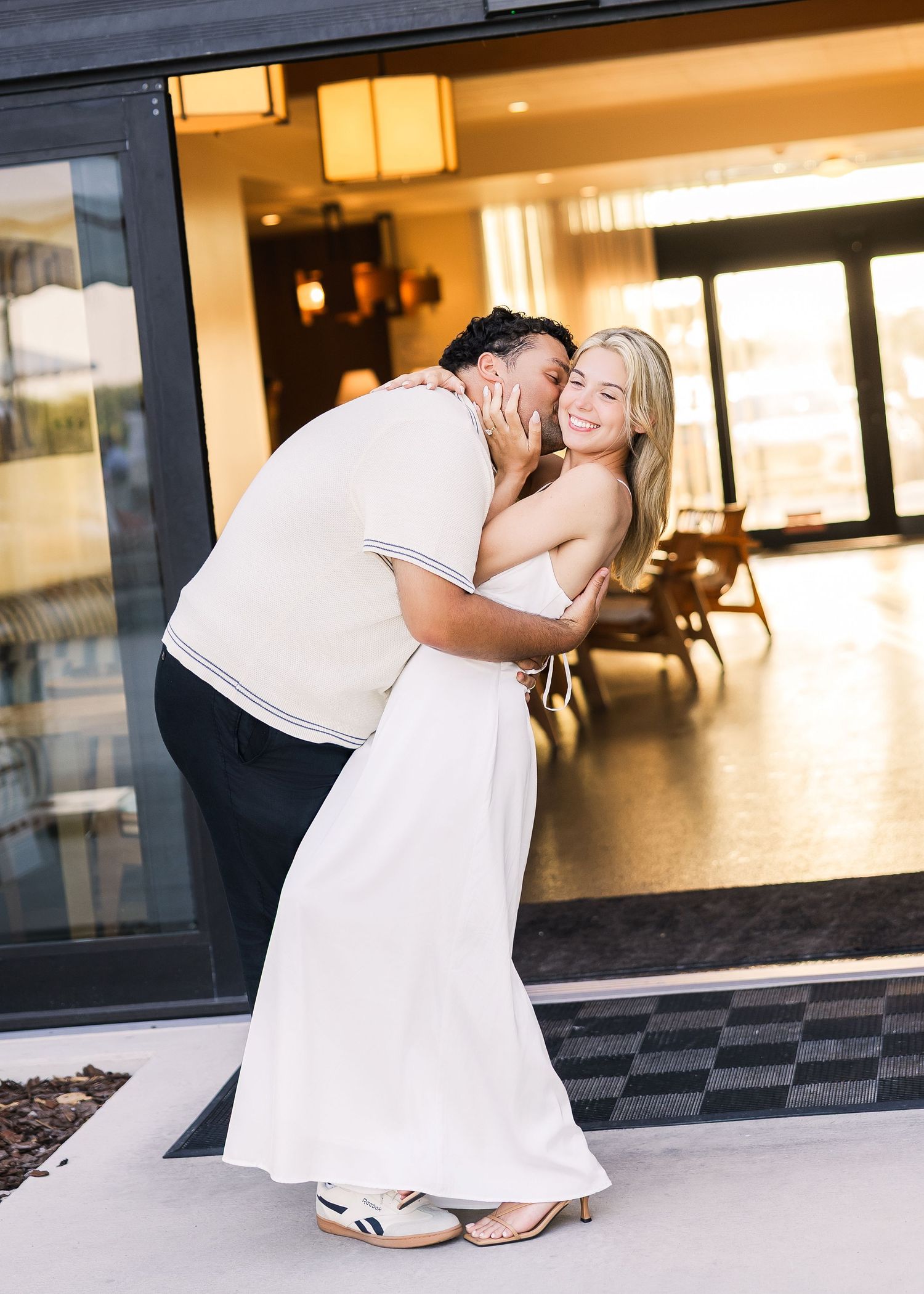 Couple embracing and kissing outside a modern building, woman in white dress smiling.