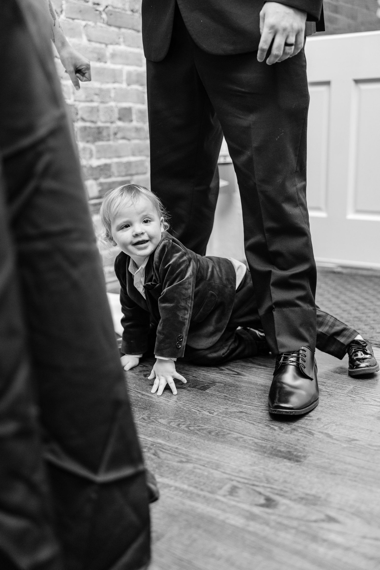 Playful black and white photo of a small child crawling between formal dress shoes on hardwood floor wedding groom bride wilmington North