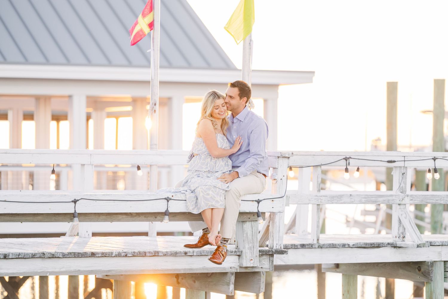 A couple sits together on a white wooden pier during golden hour.