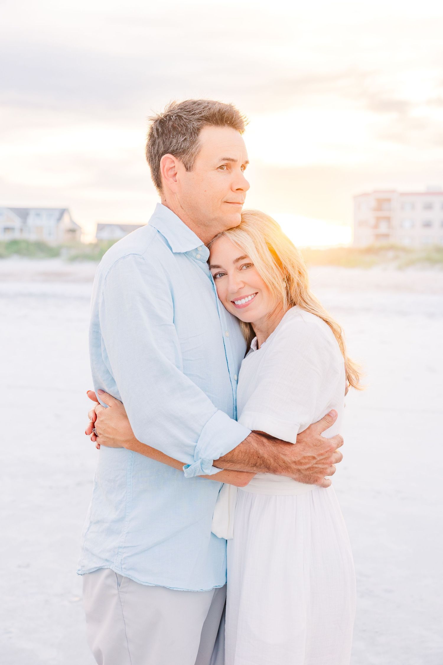 Couple embracing on a sunny beach with soft lighting and coastal homes in background.