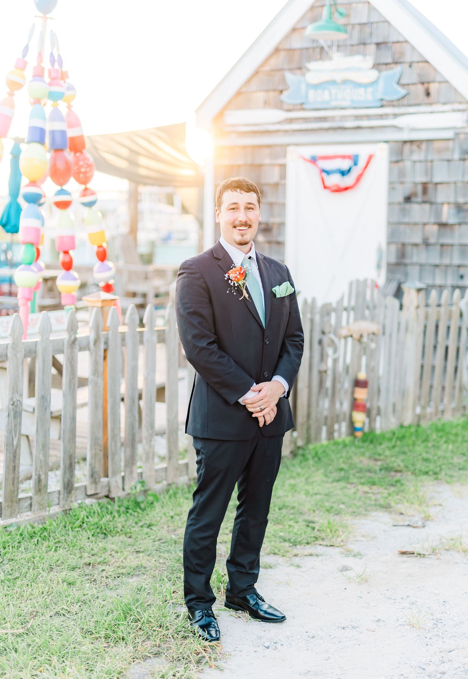 A groom in a black suit stands smiling in front of colorful decorations and a rustic building.