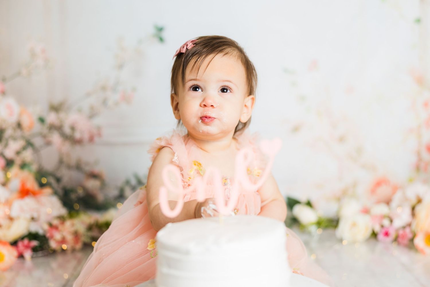 Baby girl in pink tulle dress against floral backdrop during milestone photo session.