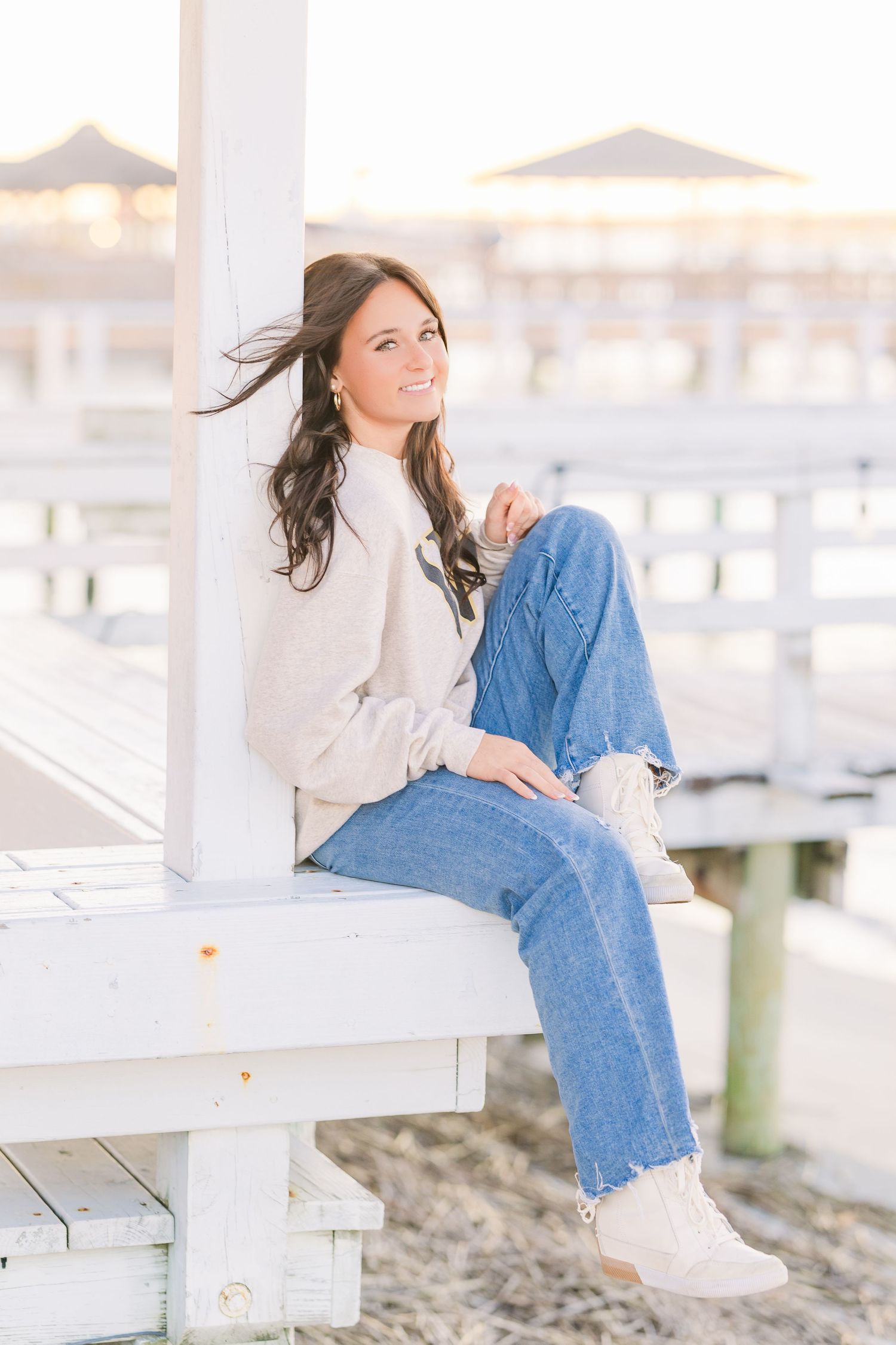 Woman in casual denim and white sweater relaxes on coastal dock during photoshoot.